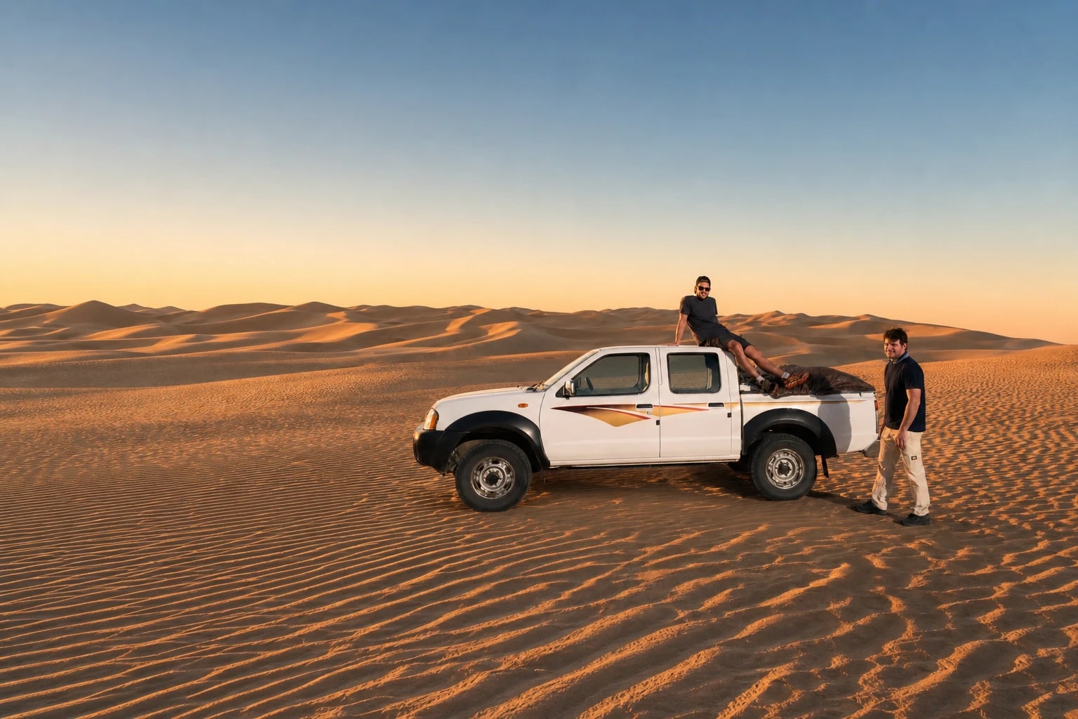 Two team members with a white pickup truck on sand dunes at sunset